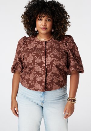 Femme aux cheveux bouclés portant un haut marron à manches bouffantes florales, un jean bleu clair, des boucles d'oreilles roses et des bracelets à son poignet droit.