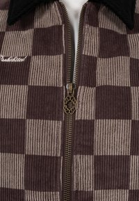 Gros plan sur une veste en velours côtelé à carreaux marron et beige avec une fermeture éclair en bronze et une broderie "Prohibited" sur le côté gauche de la poitrine.