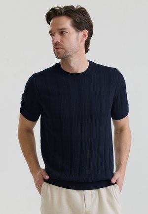 Mann mit braunen Haaren, der ein marineblaues kurzärmeliges strukturiertes Strickshirt und beige Hosen trägt, steht mit den Händen in den Taschen.