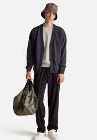 Tmavě šedý cardigan s knoflíky, světle šedá mikina, černé kalhoty a bílé tenisky. Drží olivově zelenou tašku přes rameno s černými akcenty.