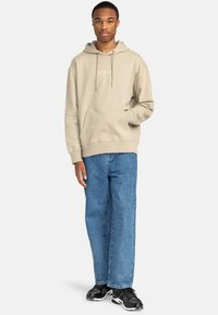 Beige hættetrøje med kængurulomme og snøre, parret med løstsiddende blå jeans og sorte sneakers. Simpelt design.
