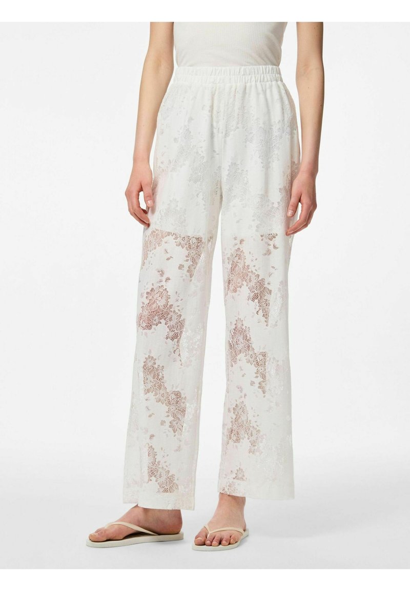 Pantalon blanc à jambes larges avec motifs en dentelle florale beige, taille élastique, porté avec un haut sans manches et des tongs beiges sur un fond uni.