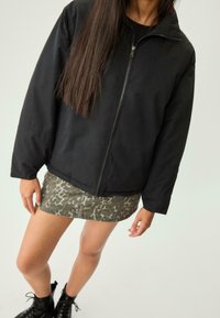 Donna che indossa una giacca nera con zip, una mini gonna a stampa leo e stivali neri stringati, in piedi davanti a uno sfondo chiaro e uniforme.