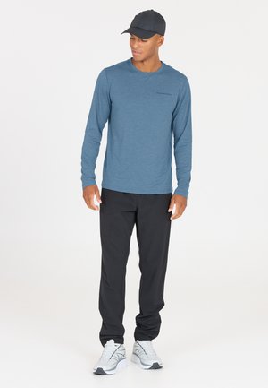 Blauwe herenlangemouwen shirt, zwarte broek, grijze sportschoenen. Shirt heeft een ronde hals; de broek is eenvoudig en ruimvallend.