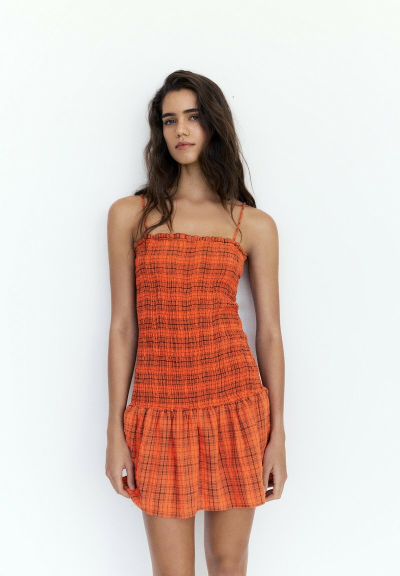 Jeune femme aux longs cheveux foncés portant une robe sans manches à carreaux orange, debout devant un fond blanc uni.