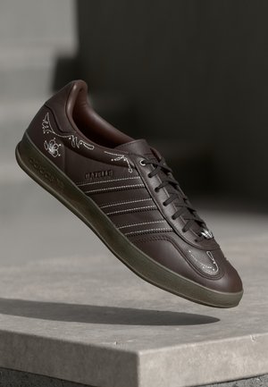 Mørkebrune Adidas Gazelle sneakers med hvidt blomstret syning og grå sål, vist svævende over en betonoverflade.