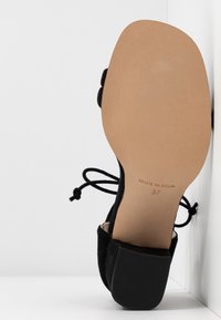 Chaussure en suede noire avec un bout arrondi, de fines lanières à la cheville et un petit talon carré. La semelle est beige avec "FABRIQUÉ EN ESPAGNE" et la taille 37 imprimée.