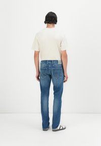 Blugi din denim albastru cu o croială dreaptă, ușor decolorati și două buzunare la spate cu detalii de cusătură. Asortați cu un tricou bej deschis.
