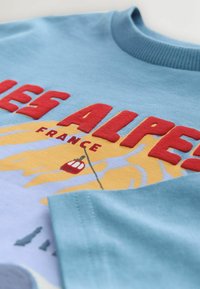 Błędny niebieski sweatshirt z haftowanym napisem "ALPES FRANCE" w czerwonym kolorze oraz graficznym wzorem przedstawiającym gondolę i górski krajobraz.