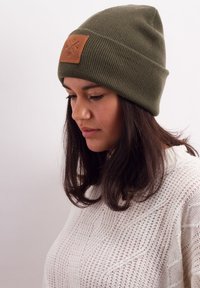 Olive green beanie made of ribbed knit fabric, featuring a brown leather patch with logo, snug fit, and fold-up brim.