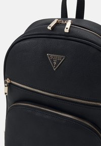 Zaino in finta pelle nera con due scomparti con zip. Il design è accentuato da hardware color oro e da un logo triangolare Guess.