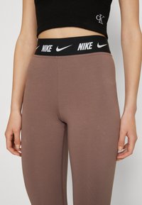 Smeđe atletske tajice s crnim pojasom s Nike logotipom, s glatkom teksturom i prilagođenim dizajnom, pogodne za aktivno nošenje.
