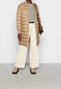 Béžová prošívaná dlouhá bunda s žebrovanými manžetami, kombinovaná s proužkovanou košilí, krémovými kalhotami s širokými nohavicemi a dvoubarevnými kotníkovými botami. Černá kabelka jako doplněk.