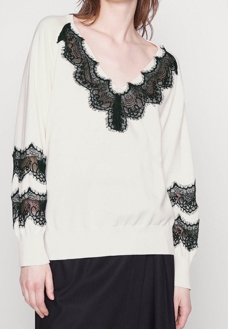 Cremfarbener Pullover mit tiefem V-Ausschnitt, schwarzen Spitzeakzenten an Ausschnitt und Ärmeln sowie einem gerippten Saum. Weiche Textur, lockerer Schnitt.