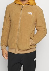 Béžová fleecová bunda na zip s kapucí, žebrovanými manžetami a lemem, s bílým logem na hrudi a bočními kapsami.