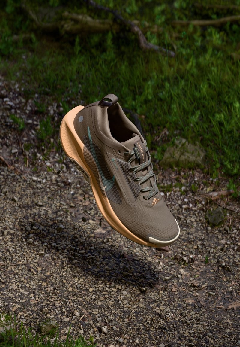 Chaussure de course grise et orange suspendue au-dessus d'un sentier de gravier avec de l'herbe verte et des arbres en arrière-plan.
