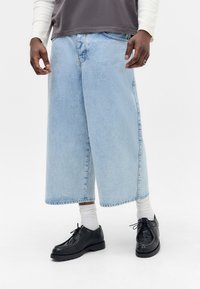 Pantalones cortos de mezclilla anchos en azul claro con un dobladillo deshilachado, combinados con zapatos de cuero negros y calcetines blancos, con una superficie texturizada.