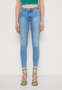 Světle modré skinny džíny vyrobené z denimu, s pěti kapsami, poutky na opasek a mírným vyblednutím. Na sobě má zelené sandály na popruzích.