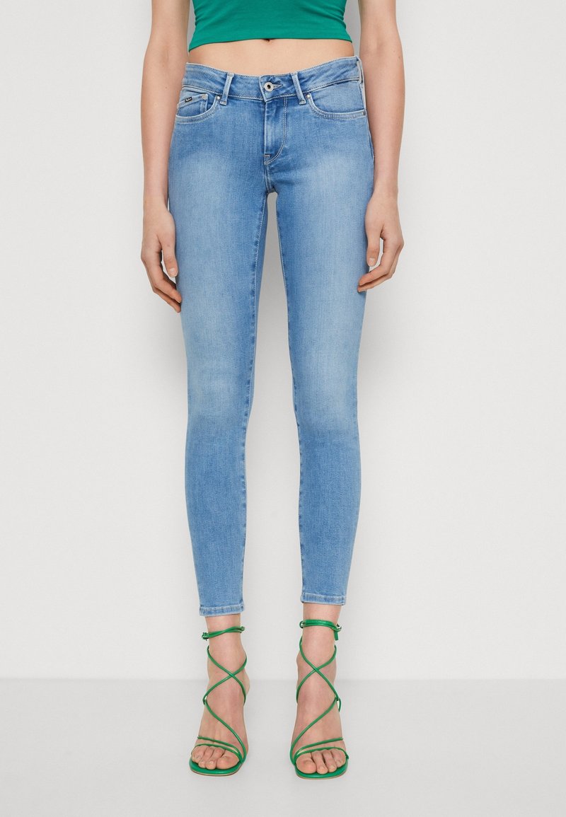 Světle modré skinny džíny vyrobené z denimu, s pěti kapsami, poutky na opasek a mírným vyblednutím. Na sobě má zelené sandály na popruzích.