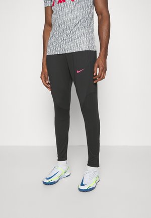 Homme portant un jogging Nike noir avec le logo Swoosh rose, une chemise blanche à motifs, et des baskets Nike blanches avec des accents bleu et vert fluo.