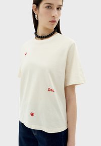 Crème katoenen t-shirt met rode geborduurde accenten "S O L" en decoratieve elementen. Korte mouwen en een ontspannen pasvorm.