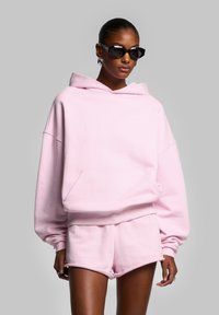 Felpa oversize rosa chiaro con tasca frontale e pantaloni corti coordinati, caratterizzata da una texture morbida e un sottile dettaglio del logo. Occhiali da sole neri come accessorio.