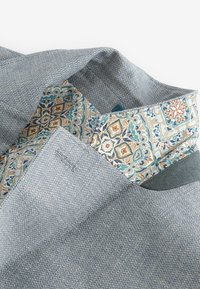 Teksturowana niebieska tkanina z geometrycznym wzorem, z detalicznym akcentem roślinnym w odcieniach beżu, zieleni i pomarańczy na wewnętrznej stronie kołnierza.