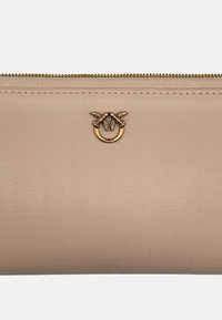 Beige leren portemonnee met een goudkleurige ringaccent met bladdetails, ritssluiting aan de bovenkant en een textuuroppervlak.