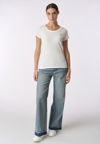 T-shirt bianca a maniche corte con scollatura rotonda e dettagli con logo, abbinata a jeans a gamba larga blu chiaro e sneakers bianche.