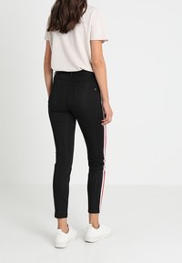 Schwarze Skinny-Jeans mit roten und weißen Seitenstreifen, hoher Taille und standardmäßigen Gesäßtaschen; kombiniert mit einem hellen T-Shirt mit kurzen Ärmeln.