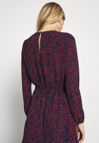 Robe bleu marine avec motif floral rouge, manches longues, détail dos en forme de goutte, et taille froncée, confectionnée en tissu doux.