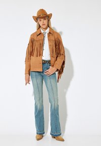 Ruskea mokkaherne-tyylinen cowboy-takki, jossa on franjeja, valkoinen kauluspaita, siniset leveät farkut ja ruskea cowboy-hattu. Huomattava vyö koristeellisella soljella.