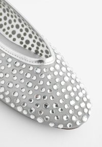 Chaussure plate argentée à bout arrondi, recouverte de strass transparents de tailles variées et doublure intérieure à pois verts sur tissu blanc.