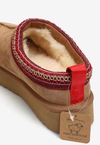 Kapcie z owczej skóry z cholewką z brązowego zamszu, ozdobione czerwoną i beżową tkana lamówką, z miękkim wnętrzem oraz czerwonym materiałowym uchwytem.