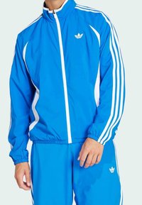 Blå träningsjacka i lättviktigt material, med vita ränder och dragkedja fram. Har en hög krage samt elastiska ärmslut och fåll.