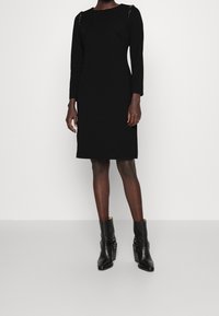Černé dlouhé šaty s rovnými liniemi, ozdobené detaily na ramenou. V kombinaci s černými vysokými botami na kotníku.