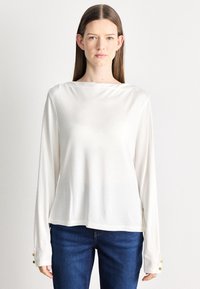 Hosszú ujjú fehér blúz, sima, könnyű anyagból. Hajónyakkal és arany gombokkal a mandzsettákon. Kék farmerrel párosítva.