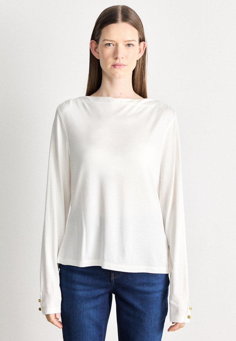 Hosszú ujjú fehér blúz, sima, könnyű anyagból. Hajónyakkal és arany gombokkal a mandzsettákon. Kék farmerrel párosítva.