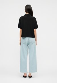 Γυναίκα με μακριά σκούρα μαλλιά φοράει μαύρο μπλουζάκι με κοντά μανίκια, ανοιχτό μπλε τζιν παντελόνι με φαρδιά πόδια και μαύρα παπούτσια με τακούνι, στέκεται με την πλάτη προς τα εμπρός.