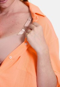 Pfirsichfarbene Bluse mit Kragen, die eine große Tasche und eine leichte Textur aufweist. Sichtbarer BH-Träger in neutralem Farbton.