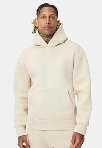 Sweat à capuche beige en matière douce, avec une poche avant et des poignets côtelés. Design simple sans motifs ni accents visibles.