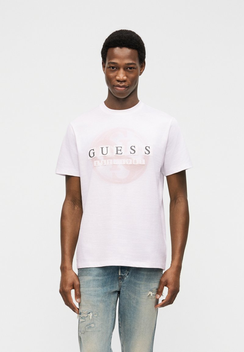 Νέος άνδρας που φοράει ένα λευκό T-shirt Guess με το λογότυπο και ανοικτόχρωμα σκισμένα τζιν, στέκεται μπροστά σε ένα απλό ανοιχτό γκρι φόντο.