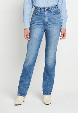 Person, die hoch taillierte, gerade geschnittene blaue Jeans, ein hellblaues, eingestecktes Hemd mit Knöpfen und weiße, hochhackige Stiefeletten vor weißem Hintergrund trägt.