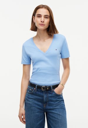 Femme aux cheveux bruns raides, portant un T-shirt bleu clair à col en V, un jean bleu, une ceinture noire et un bracelet en or au poignet gauche.