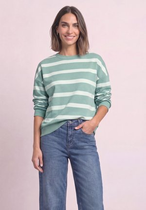 Kvinde smiler iført en grøn og hvid stribet langærmet sweater og blå jeans med den ene hånd i lommen mod en lyserød baggrund.