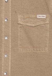 Beige denimskjorte med frontlukking med knapper, sølvfargede trykknapper og en lomme på venstre bryst med en rød og hvit Calvin Klein-etikett.