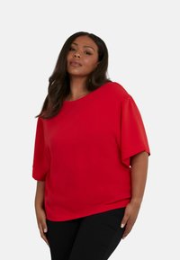 Top rosso a maniche corte con un taglio morbido, scollatura rotonda e texture liscia. Indossato con pantaloni neri, che evidenziano le caratteristiche di design semplici.