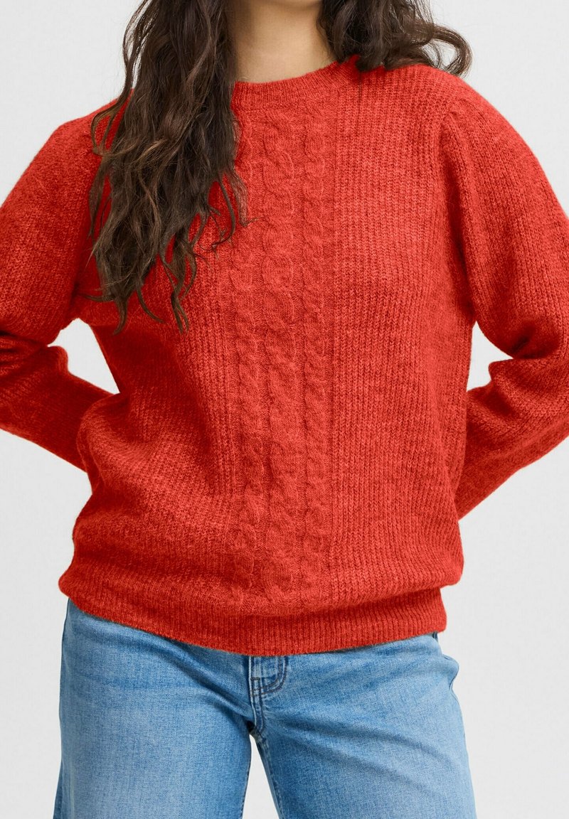 Pull rouge en tricot avec un motif torsadé texturé sur le devant, col rond et manches larges, associé à un jean bleu clair.