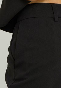 Pantalones negros hechos a medida de una tela suave, con delantero plano y bolsillo, acabado limpio y detalle de costura sutil.