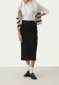 Nainen, jolla on päällään valkoinen paita, musta korkeavyötäröinen midihame, jossa on etureikä, mustat loafersit, kermaiset sukat ja kerma-collegepaita heitettynä hartioiden ylle.